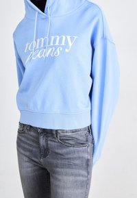 Sudadera azul clara con el logo blanco de "Tommy Jeans", capucha y puños acanalados, combinada con jeans grises que presentan un desvanecimiento sutil.