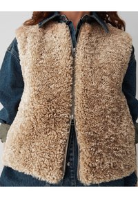 Gilet en fausse fourrure beige avec une fermeture éclair à l'avant, surface texturée et ourlet arrondi, superposé sur une chemise en denim foncé.
