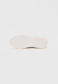 Skoens yttersåle i hvit gummi, har mønstret grep med små sirkulære elementer. Glatt overflate, minimalistisk design og flat profil.