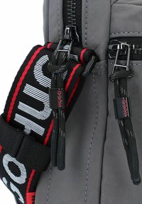 Mochila de tela gris con correas ajustables en negro y rojo con el texto "HUGO". Cierres de cremallera y acentos duraderos visibles.