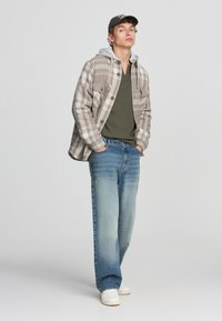 Szara w kratkę flanelowa koszula z kapturem, oliwkowa koszulka pod spodem, jasnoniebieskie dżinsy i białe trampki. Model stoi z dłońmi w kieszeniach.