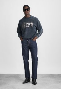 Tumehall sweatshirt metallilise "KISS" logoga, koos tumesiniste teksade ja mustade pahkluupakkudega. Modell kannab musti päikeseprille.