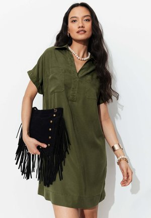 Femme portant une robe chemise vert olive, tenant une pochette noire à franges, accessoirisée d’un collier de perles, de bracelets et d’un bracelet manchette en or.