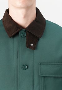 Grøn jakke med en brun kordfløjskrave, knaplukninger og en brystlomme med stikdetaljer. Glat og struktureret stof.