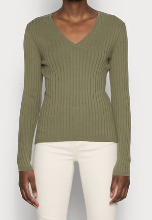 Personne portant un pull côtelé vert olive à col en V à manches longues et un pantalon beige clair, debout devant un fond neutre.