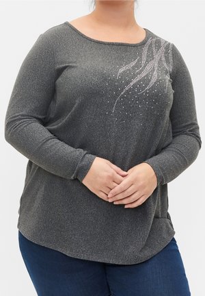 Vrouw draagt een donkergrijs shirt met lange mouwen en een ruitsteendesign in de vorm van een krul op de borst, handen in elkaar gevouwen, gecombineerd met donkerblauwe jeans.