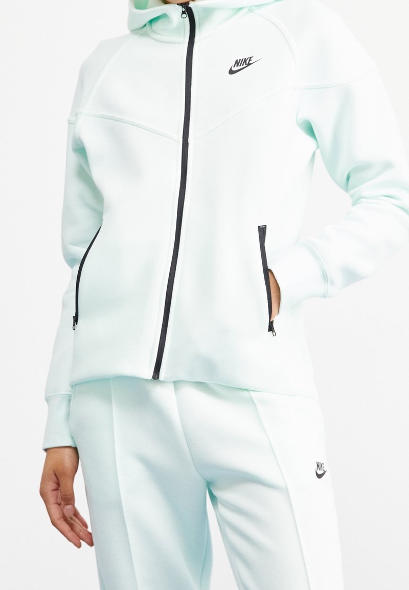 Light mint green Nike zip-up jacket with a hood, featuring black zipper and pockets, soft material, and a sleek, structured design.