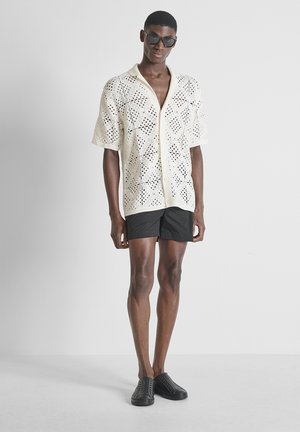 Man draagt zwarte zonnebril, witte opengebreide overhemden met korte mouwen en knopen, zwarte korte broek en zwarte instapschoenen, staand tegen een effen achtergrond.