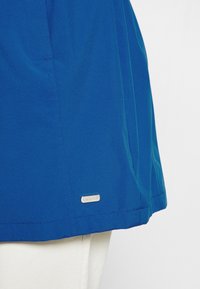 Panneau en tissu bleu avec des coutures cousues et une petite étiquette métallique rectangulaire de marque près du bord inférieur, sur fond textile blanc.