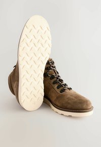 Botín de ante marrón con una suela texturizada color crema. Presenta ojales de metal y cordones oscuros. Destaca por un diseño robusto pero flexible.