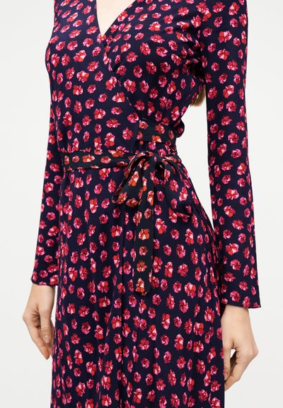Μπλε σκούρο φόρεμα τύπου wrap με κόκκινο και ροζ ανθισμένο σχέδιο, μακριά μανίκια και ζώνη στη μέση δεμένη, φορεμένο από μια γυναίκα με τα χέρια χαλαρά δίπλα στα πλευρά.
