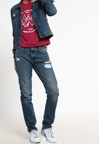 Denim takki, viininpunainen huppari graafisella printillä, kuluneet siniset farkut ja valkoiset lenkkarit. Rentoa asua erilaisilla tekstuureilla ja väreillä.