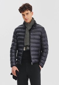 Czarna kurtka ocieplana z zapięciem na zamek, z poziomym pikowanym wzorem. W połączeniu z czarno-białym wzorzystym szalikiem. Widoczne czarne spodnie.