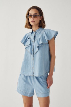 Žena oblečená v svetlomodrom bezrukávovom rifľovom top s gombíkmi a volánikmi na ramenách a v rifľových kraťasoch, doplnená o hnedé slnečné okuliare.