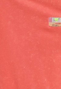 Tissu couleur corail avec une texture douce et mate ; présente une étiquette vintage dans le coin supérieur droit avec un cœur vert et des accents rayés.