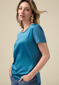 Tricou teal cu mânecă scurtă, cu o textură fină și guler rotund, asortat cu blugi de culoare albastru deschis. Accesoriile includ cercei mari din aur.