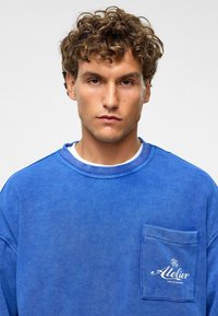 Blauwe sweater met ronde hals van katoen, beschikt over een voorvak met geborduurde tekst. Zachte textuur, losse pasvorm en minimalistisch ontwerp.