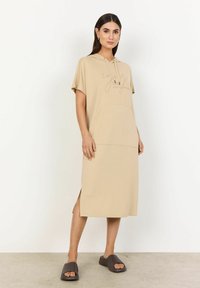Beige huvtröja med korta ärmar, med en framficka och sidoventilation. Modellen bär bruna slidesandaler.