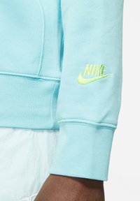 Felpa azzurra con una texture morbida, caratterizzata da un logo Nike neon giallo ricamato sul polsino sinistro.