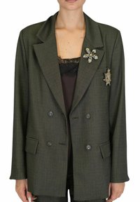 Blazer doppiopetto verde scuro in tessuto strutturato con rever a scollo, con due spille decorative sul lato sinistro.