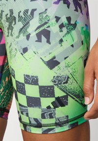 Mnohobarevné sportovní šortky s geometrickým vzorem v zelené a černé barvě, s dynamickými tvary, hladkou texturou a logem Adidas.