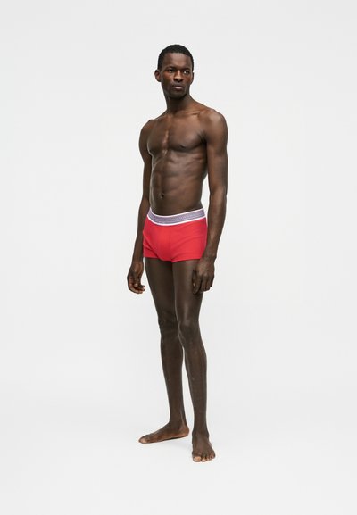 Homme athlétique à la peau foncée, debout pieds nus, portant un boxer rouge avec une ceinture blanche à motifs, sur un fond gris uni.