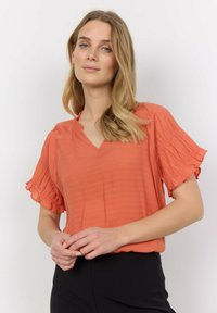 V-neck, short-sleeved blouse in textured orange fabric. Features ruffled sleeves and a relaxed fit. Paired with fitted black pants.