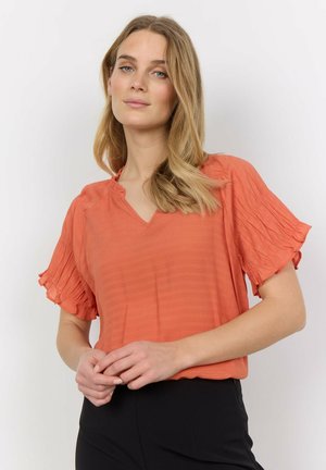 V-neck, short-sleeved blouse in textured orange fabric. Features ruffled sleeves and a relaxed fit. Paired with fitted black pants.