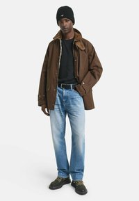 Bruine buitenjas met een corduroy kraag, zwart shirt eronder, lichtblauwe jeans en een zwarte gebreide beanie, afgerond met groene en zwarte laarzen.