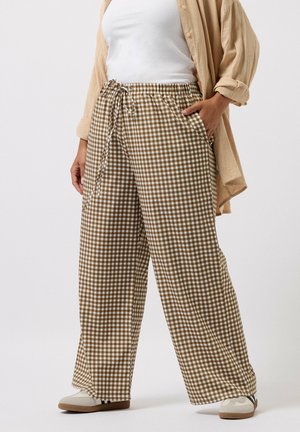 Osoba ubrana w beżowe i białe, szerokie spodnie w kratę, biały top na ramiączkach, beżową otwartą koszulę oraz jasne sneakersy, stojąca z jedną ręką w kieszeni.