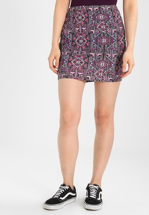 Mujer con una falda corta con un patrón geométrico colorido y zapatillas casuales en blanco y negro, de pie frente a un fondo sencillo.