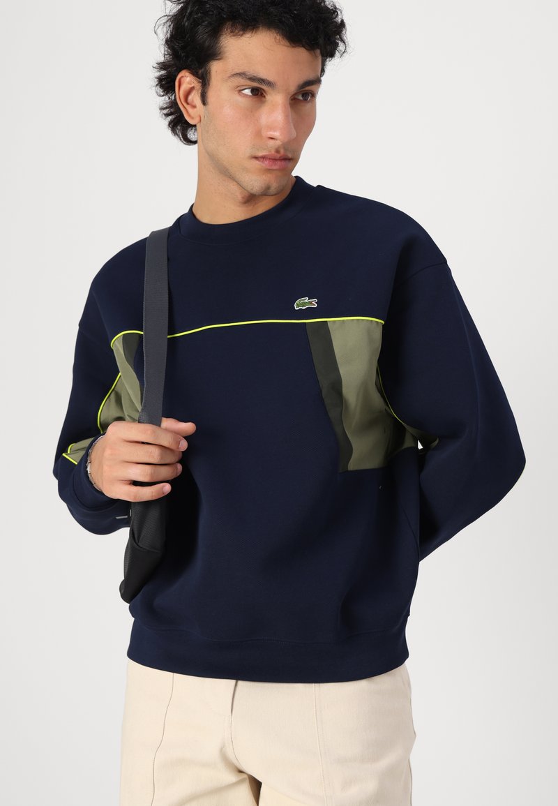 Sweatshirt marine avec des accents verts et une bande jaune, doté d'un col rond et d'une texture douce. Logo Lacoste sur la poitrine.