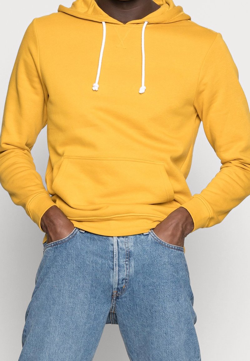 Persona con una sudadera amarilla mostaza con cordones blancos y pantalones vaqueros azules, con las manos metidas en los bolsillos delanteros.
