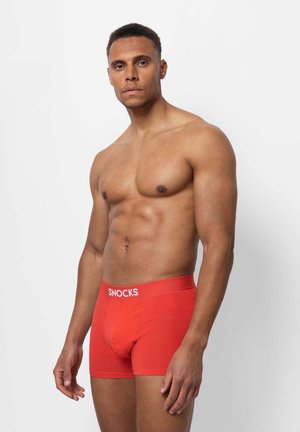 Shorts de boxeur rouges en tissu doux, offrant une coupe ajustée et un logo blanc "SNOCKS" sur la ceinture. Design minimaliste, texture lisse.