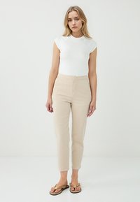 Een vrouw staat tegen een witte achtergrond, gekleed in een strakke witte top en beige broek, in combinatie met casual sandalen.