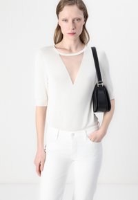 Un haut en tricot blanc avec un décolleté en V profond et des manches courtes, associé à un jean blanc. Un petit sac à main noir repose sur l'épaule.
