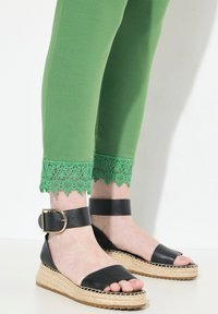 Leggings verdes con ribete de encaje floral en los tobillos, combinados con sandalias de esparto negras que cuentan con una tira ancha y hebilla en el tobillo.