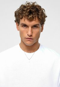 Witte T-shirt met een ronde hals, gedragen door een persoon met krullend haar, met een zilveren ketting met een kleine hanger.