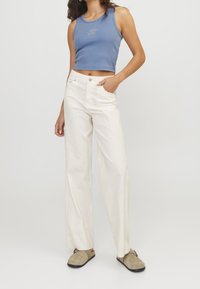 Modrá vroubkovaná crop top s malou grafikou, doplněná o bílé kalhoty s širokými nohavicemi. Modelka má na sobě béžové nazouvací boty. Neutrální pozadí.