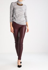 Frau in einem grauen Rollkragenpullover, bordeauxroten Lederhosen, schwarzen spitzen Ballerinas und einer großen bernsteinfarbenen Statement-Halskette vor einem schlichten Hintergrund.