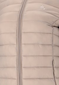 Lichtbeige pufferjack met verticale stiksels, voorzien van een volledige ritssluiting en een klein logo aan de bovenkant links. Zachte, gladde textuur.