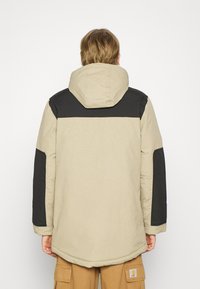 Beige og svart hetteparka med en teksturert overflate, med solid fargeblokkering og elastiske mansjetter. Baksidevisning viser en avslappet passform.