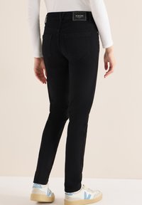 Vaqueros pitillo de mezclilla negra con cintura de altura media, bolsillos traseros y una etiqueta lisa en la cinturilla. Combinados con zapatillas blancas con detalles en azul.