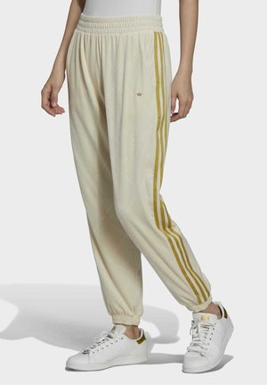 Persoon draagt crèmekleurige joggingbroek met gouden strepen en klein logo, gecombineerd met witte sneakers met gouden details.