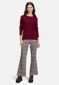 En kvinna iklädd en vinröd tröja, rutiga utsvängda byxor och mörka klackboots, stående mot en ljus enfärgad bakgrund.