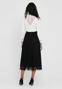 Haut blanc à manches longues avec un dos en dentelle associé à une jupe plissée noire. Des escarpins noirs complètent la tenue.