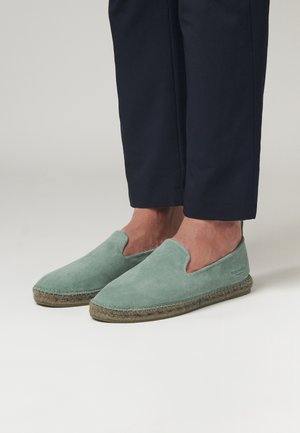 Zapatos verdes de ante sin plantilla con una suela de yute trenzada al estilo espadrille, que presentan una punta redondeada y un diseño con el talón abierto.