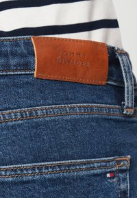 Καφέ δερμάτινη ετικέτα με το λογότυπο «Tommy Hilfiger» ραμμένη στην τσέπη από μπλε τζιν, άτομο που φοράει λευκό και ναυτικό ριγέ πουκάμισο.