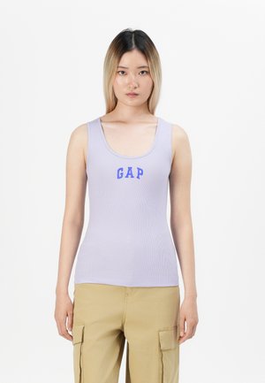 Ung kvinne med langt ombre-hår iført en lavendel tanktopp med "GAP"-logo og beige cargo-bukser, stående mot hvit bakgrunn.