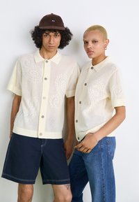 Strikkede, kremfargede t-skjorter med kort erme og paisley-mønster, med knappet krage, kombinert med denim og mørke shorts. Brun caps med logo.